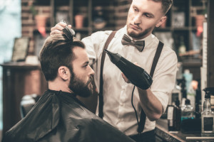 New hairstyle. Side view of young bearded man getting groomed at hairdresser with hair dryer while sitting in chair at barbershop
